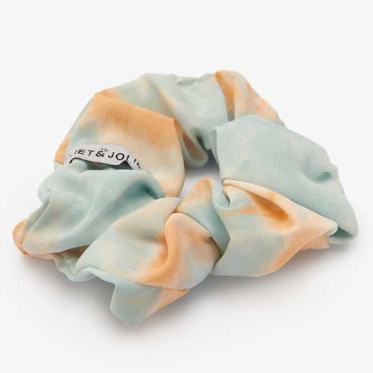 Bad hair day? Geen probleem want met deze vrolijke tie-dye scrunchie maak je van ieder kapsel een feestje!   De scrunchie is gemaakt van polyester en heeft een diameter van ongeveer 13cm. De stof heeft een blauw met licht oranje tie-dye effect.